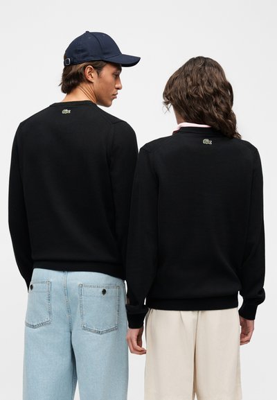 Deux personnes vues de dos portant des hauts à manches longues noirs avec de petits logos verts sous les cols ; l'une en jean bleu clair et casquette marine, l'autre en short beige.