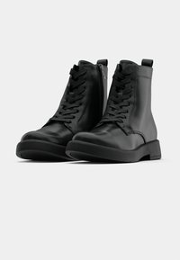Bottes en cuir noir à lacets, avec fermetures éclair latérales et semelles épaisses en caoutchouc, posées sur un fond blanc.