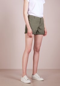 Pantaloni corti in cotone verde oliva con risvolti, tasche laterali e chiusura con bottone frontale, abbinati a sneakers bianche e una t-shirt bianca semplice.