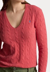 Koralowy sweter dziany z dekoltem w serek i wzorem warkoczowym. Na piersi małe niebieskie logo. Długie rękawy z prążkowanymi mankietami.