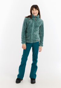 Djevojčica koja se smije u tirkiznoj mekanom zip-up jakni, tirkiznim i bijelim hlačama te crnim čizmama, stoji nasuprot bijeloj pozadini.