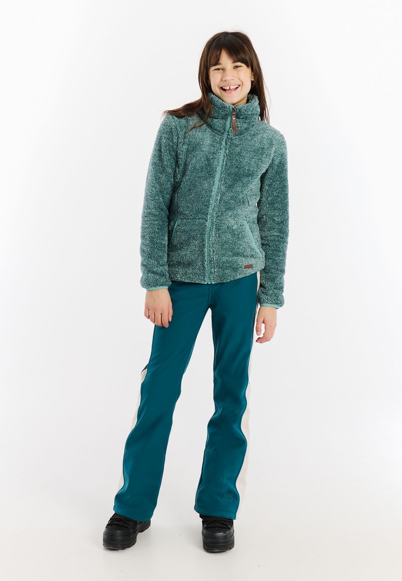 Djevojčica koja se smije u tirkiznoj mekanom zip-up jakni, tirkiznim i bijelim hlačama te crnim čizmama, stoji nasuprot bijeloj pozadini.