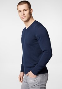 Granatowy sweter z dekoltem w serek, wykonany z materiału dzianinowego, z długimi rękawami i ściągaczami, noszony na jasnoszarych dżinsach.