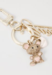 Zlatý kľúčenka s príveskom v tvare medvedíka ozdobená rhinestone kamienkami, ružovými akcentmi a malým srdiečkovým príveskom s nápisom "GUESS".