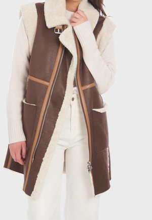 Femme portant une veste sans manches en peau lainée marron avec une doublure en fourrure crème par-dessus un pull blanc et un pantalon blanc, mains près du col et du côté de la veste.