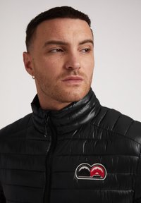 Giacca a vento nera con colletto alto, tessuto trapuntato, zip completa e un logo a doppia montagna rosso e grigio sul lato sinistro del petto.