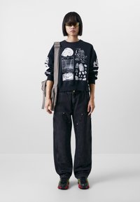 Felpa nera con grafiche bianche, abbinata a pantaloni di jeans neri larghi con dettagli gialli e una borsa a spalla grigia.