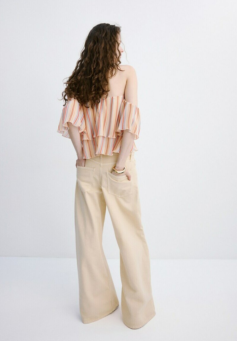 Femme aux cheveux bouclés portant un haut rayé à épaules dénudées et un pantalon ample beige, debout, les mains dans les poches arrière.