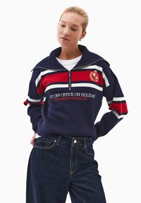 Granatowy sweter z białymi i czerwonymi paskami, wysoki kołnierz, zamek błyskawiczny do połowy oraz haftowany tekst. W zestawie z ciemnoniebieskimi dżinsami.