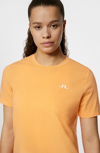 Orange bomullströja med rundhals, korta ärmar och en liten vit logotyp på vänster bröst. Slät textur, avslappnad passform.