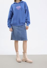 Sudadera con capucha azul con texto gráfico rosa, combinada con una falda de denim y botas de tobillo glossy rosa claro con suelas gruesas.