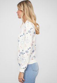 Sweatshirt à motif floral en blanc, avec des fleurs bleues et roses. Manches et ourlet côtelés, avec une coupe décontractée. Porté avec un jean bleu clair.
