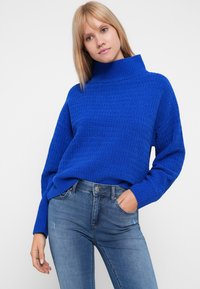 Pull en maille bleu vif à col montant avec motif texturé, coupe décontractée et manches longues, associé à un jean en denim bleu clair.