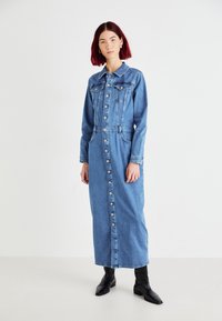 Denimkjole med lange ermer, knapper foran, to brystlommer og en figursydd midje. Stoffet er lyseblått, og den rekker nedenfor anklene.