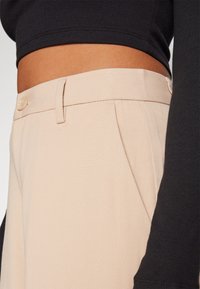 Pantalons beige ajustés avec une texture lisse, dotés d'une fermeture à bouton, de poches latérales et de plis subtils. Associés à un haut noir côtelé.