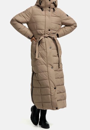 Manteau beige long et rembourré avec une ceinture nouée à la taille, capuche, boutons-pression, et bottes noires épaisses portées par une personne avec les mains sur les hanches.