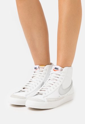 Sneakers alte Nike in pelle bianca. Presentano un logo Swoosh grigio, lacci bianchi, suola in gomma e dettagli in suede grigio chiaro sulla punta.