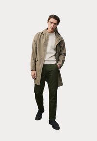 Lys khaki vandtæt jakke med hætte, parret med en cremefarvet sweater og mørkegrønne bukser. Sorte sko fuldender looket.