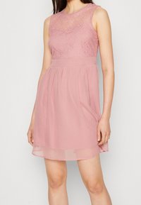 Robe rose avec un corsage en dentelle et une couche semi-transparente, sans manches, et une jupe fluide plissée. Texture douce et silhouette féminine.