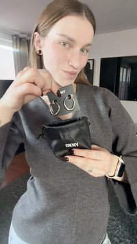 Brincos de argola prateados exibidos num cartão preto, segurado sobre um estojo preto da DKNY. O modelo veste um suéter cinza e tem verniz escuro nas unhas.