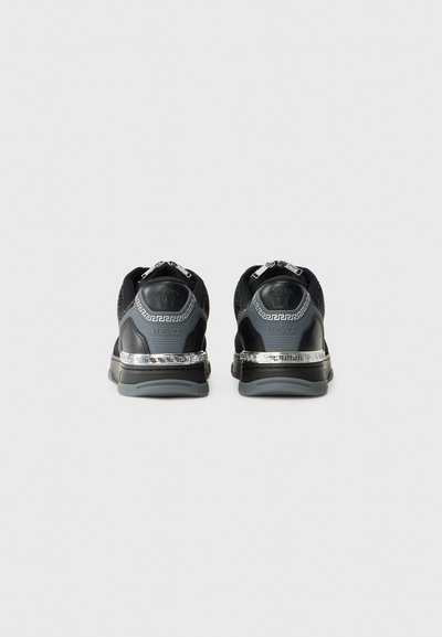 Black athletic shoes with mesh and leather uppers, featuring a silver patterned midsole and rear zip accents. Versace logo on heel.