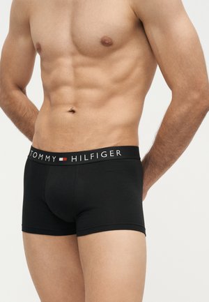 Wypoczęta męska sylwetka w czarnych bokserkach Tommy Hilfiger, dłonie spoczywają za plecami, pokazując umięśniony brzuch i kolczyk w pępku.