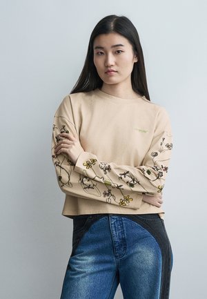 Νεαρή γυναίκα με μακριά σκούρα μαλλιά, ντυμένη με μπεζ φουτέξ με κέντημα λουλουδιών και δίχρωμα μπλε τζιν παντελόνια, σε απλό γκρι φόντο.