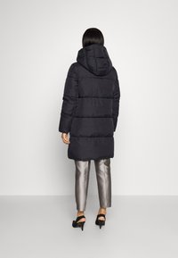 Puffjakke i sort med hætte, kraftig quiltning og en mid-length design. Båret over skinnende sølvbukser og sorte hæle.