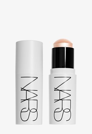 Stick de maquillage NARS blanc avec base noire à tourner, présentant un enlumineur champagne scintillant à côté de son capuchon blanc assorti.