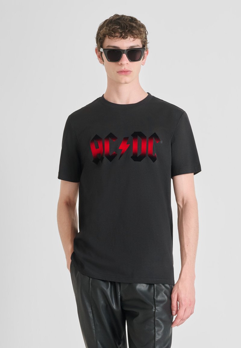 Camiseta de algodón negra con un atrevido logo de AC/DC en rojo, modelada con gafas de sol negras y combinada con pantalones de cuero sintético negros.