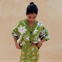 Cardigã verde oversize com padrões de olhos e corações, caracterizado por um decote em V profundo, mangas largas e botões pretos, combinado com uma saia com padrão de olhos correspondente.