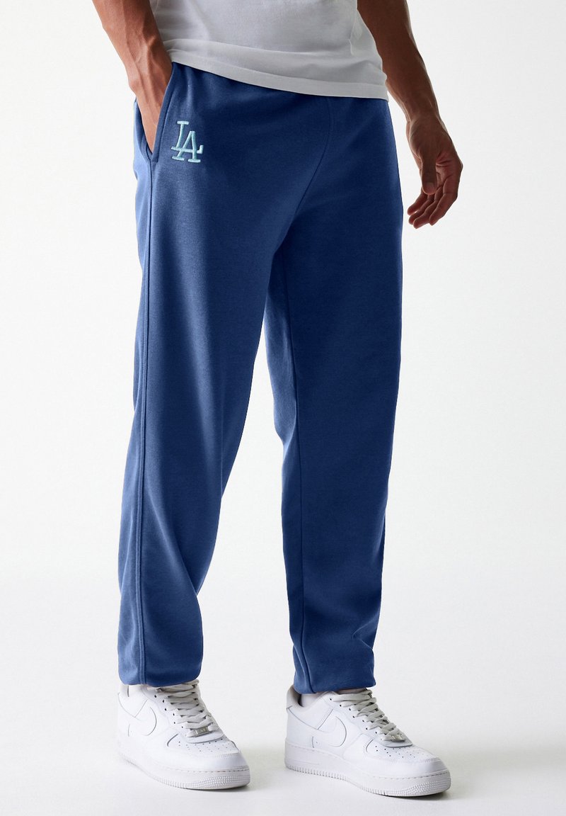 Un pantalon de survêtement bleu en tissu doux, avec un cordon de serrage blanc, des poches latérales et un logo LA bleu clair sur la hanche gauche.