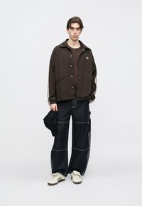 Καφέ σακάκι με κουμπιά και λευκές ρίγες, μαύρο πουκάμισο, φαρδιά σκούρα μπλε τζιν με ραφές και λευκά αθλητικά παπούτσια. Καπέλο στο χέρι.