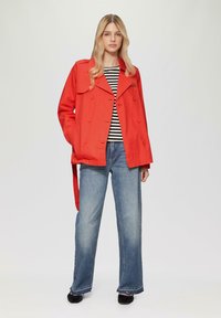 Sarkans, divkāršs tranšejas mētelis ar plecu epauletēm, uzvilkts virs melna un balta svītraina topa, kombinēts ar platām zila džinsu biksēm.