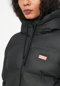 Žena nosi crnu zimske jakne s visokim ovratnikom i oznakom "Hunter" na prsima, zatvorenoj s patentnim zatvaračem, s vidljivim kapuljačom na čipku.