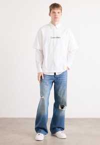 Balta trumpų rankovių marškiniai su "Calvin Klein" logotipu, uždėti ant ilgarankovių marškinių, derinami su pablukusiais mėlynais plačiomis kelnėmis su suplyšimu.