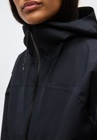 Κοντινό πλάνο ενός ατόμου που φοράει μαύρο μπουφάν με κουκούλα, με φερμουάρ στο μπροστινό μέρος και κορδόνι στην κουκούλα.