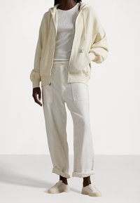 Kremowa bluza z kapturem zapinana na zamek z kieszenią z przodu, w połączeniu z kremowymi spodniami o luźnym kroju. Buty wykonane z jasnobeżowego płótna, na sznurówki.