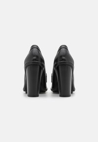 Un par de zapatos de tacón alto de cuero negro mostrados desde atrás sobre un fondo blanco.