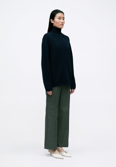 Navy coltrui, oversized pasvorm, gecombineerd met groene geruit wijde broek en witte puntige schoenen. Schoon, minimalistisch ontwerp.