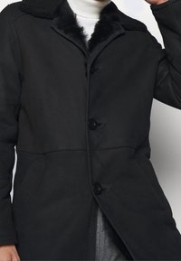 Manteau extérieur noir avec un col en fausse fourrure noire, fermeture à boutons à l'avant, deux poches latérales et une texture lisse. Présente des éléments de design sur mesure.