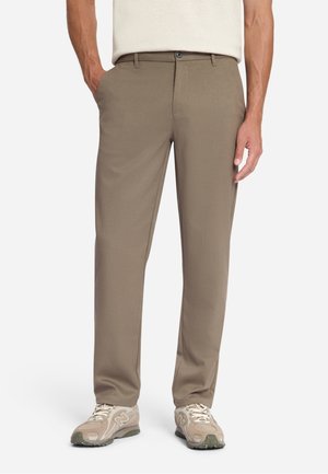 Man draagt beige rechtvallende broek met zakken, gecombineerd met bijpassende beige sneakers op een effen witte achtergrond.