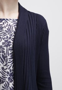 Kvinna som bär en marinblå ribbad cardigan över en vit topp med marinblå blommönster och bladmönster.