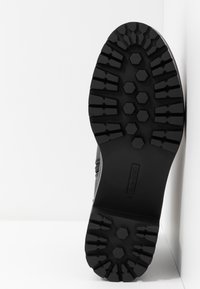 Semelle d'une botte noire avec des crampons profonds hexagonaux et linéaires, positionnée verticalement contre un mur blanc d'angle.