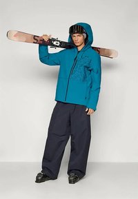 Noor mees sinises suusajakos ja mustades pükstes, hoides suuski õlal, kandes musti suusaprille ja saapaid, seismas ühtlase tausta ees.