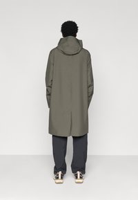 Hosszú, olívazöld, vízálló kabát kapucnival, egyenes szabással és hátsó szellőzővel, sötét nadrággal és sportcipővel párosítva.