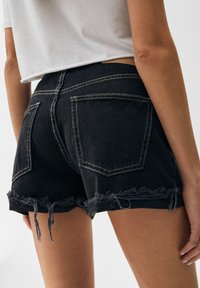 Zwarte denim shorts met rafelige zoom, contrasterende lichte stiksels en twee achterzakken. De stof heeft een gladde textuur.