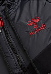 Sort polstret jakke med en fremtrædende rød broderet logo. Den har en lynlås med sort kant og teksturerede stofdetaljer.