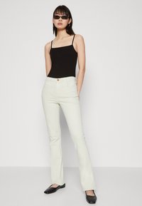 Jean évasé blanc au texture lisse, avec une fermeture à bouton sur le devant et des poches, associé à un débardeur ajusté noir et des ballerines noires.