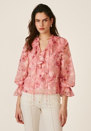 Mujer con blusa transparente rosa con estampado floral y volantes, y pantalones beige claro, de pie frente a un fondo liso.